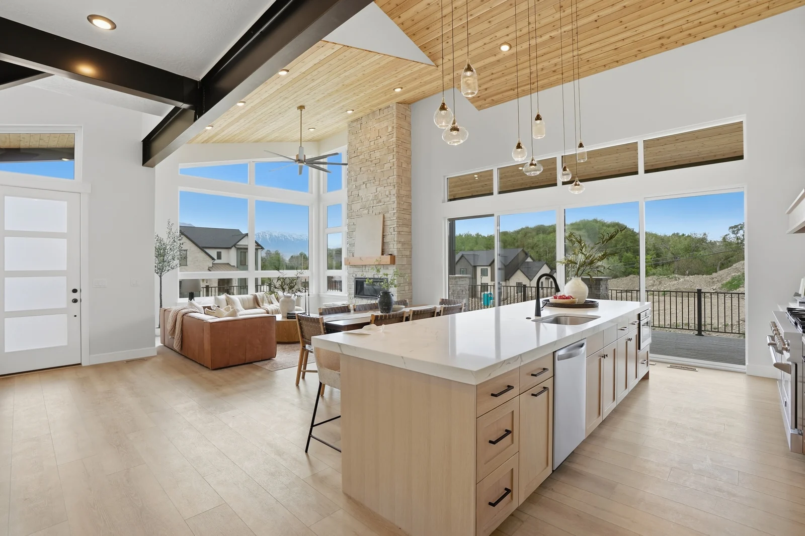 View across the kitchen island toward the window-lined great room, dining area, and exposed dark ceiling beam in the Denali model, American Fork.