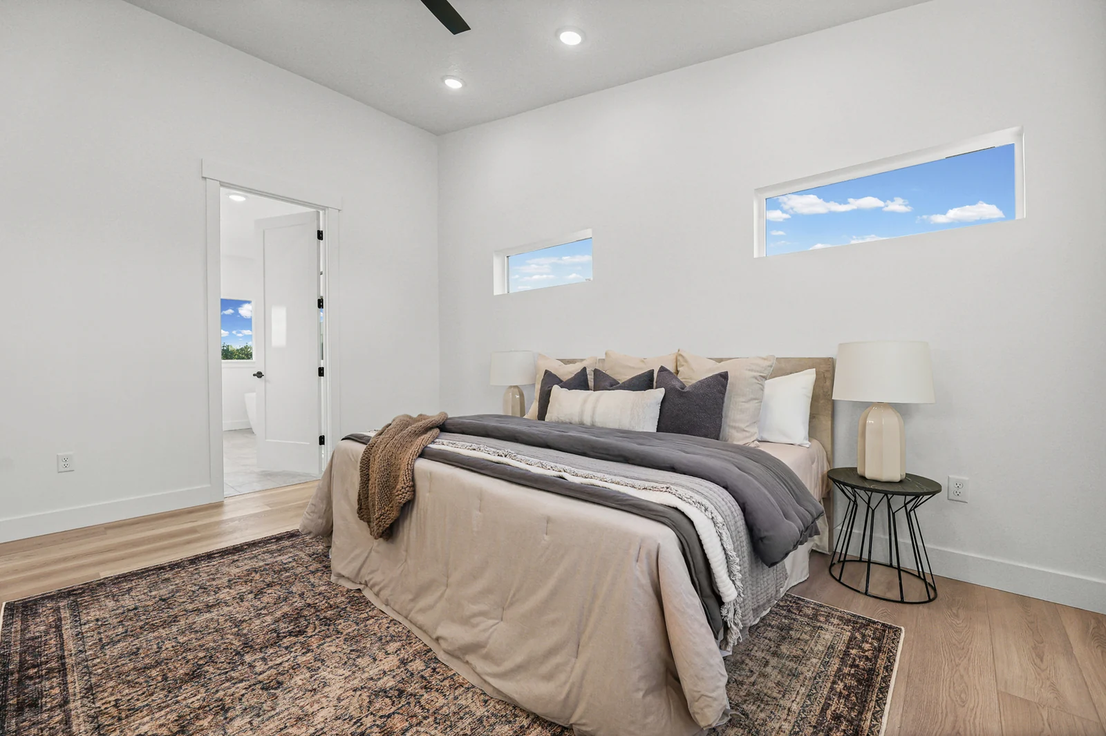 Staged bedroom features a bed, layered rug, side tables, white walls, and recessed lighting in Hawthorne Farmhouse, Ogden.