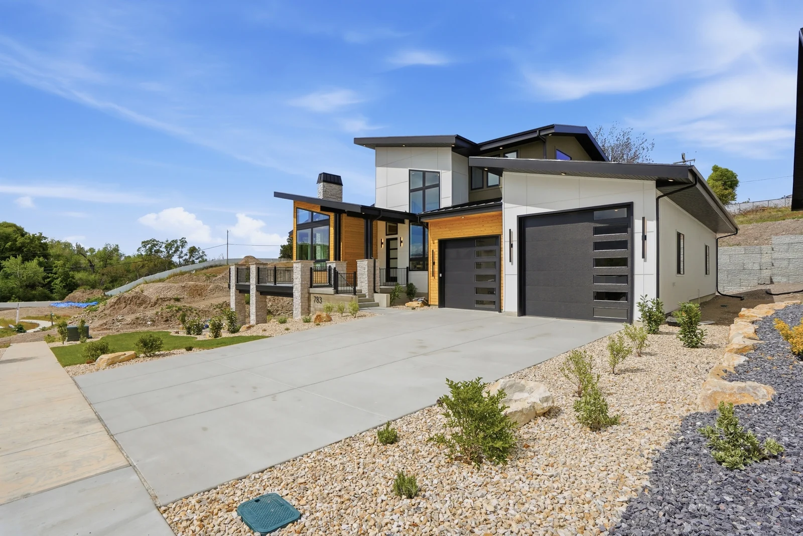Daytime exterior view from the driveway showing the modern facade, dark garage doors, concrete paving, and planted rock beds in the Denali model, American Fork.