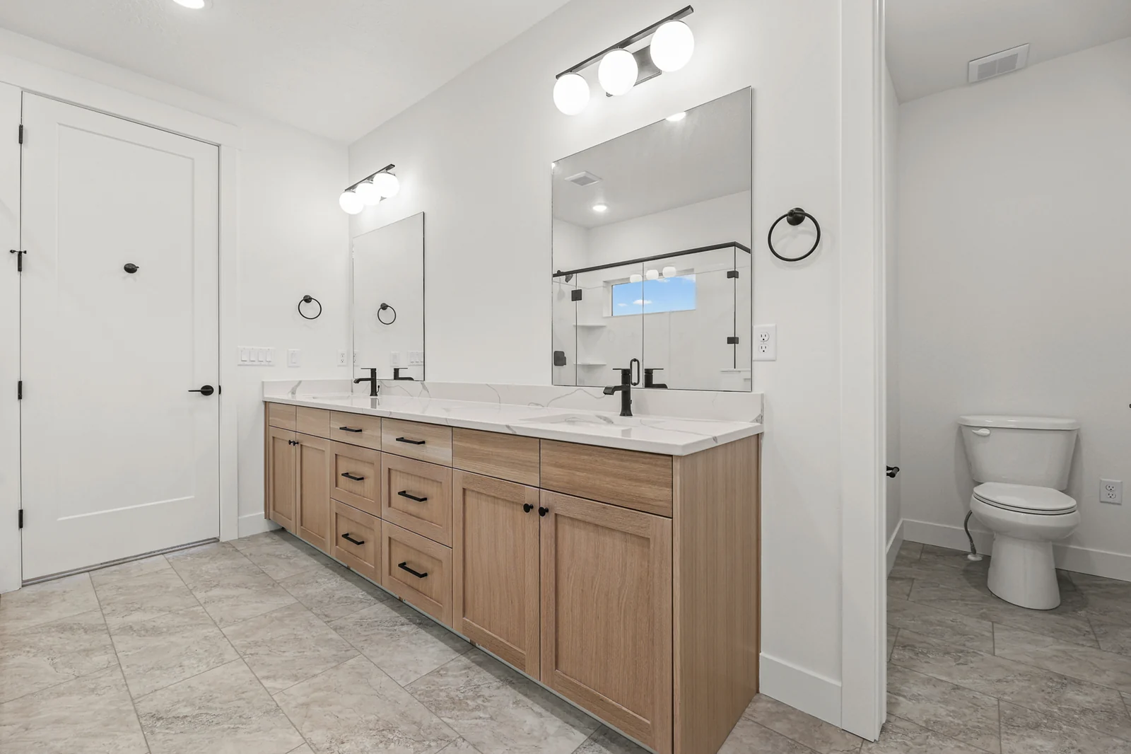Primary bathroom with wood double vanity, large mirror, toilet, black fixtures, and light tile floor in Hawthorne Modern, Ogden.