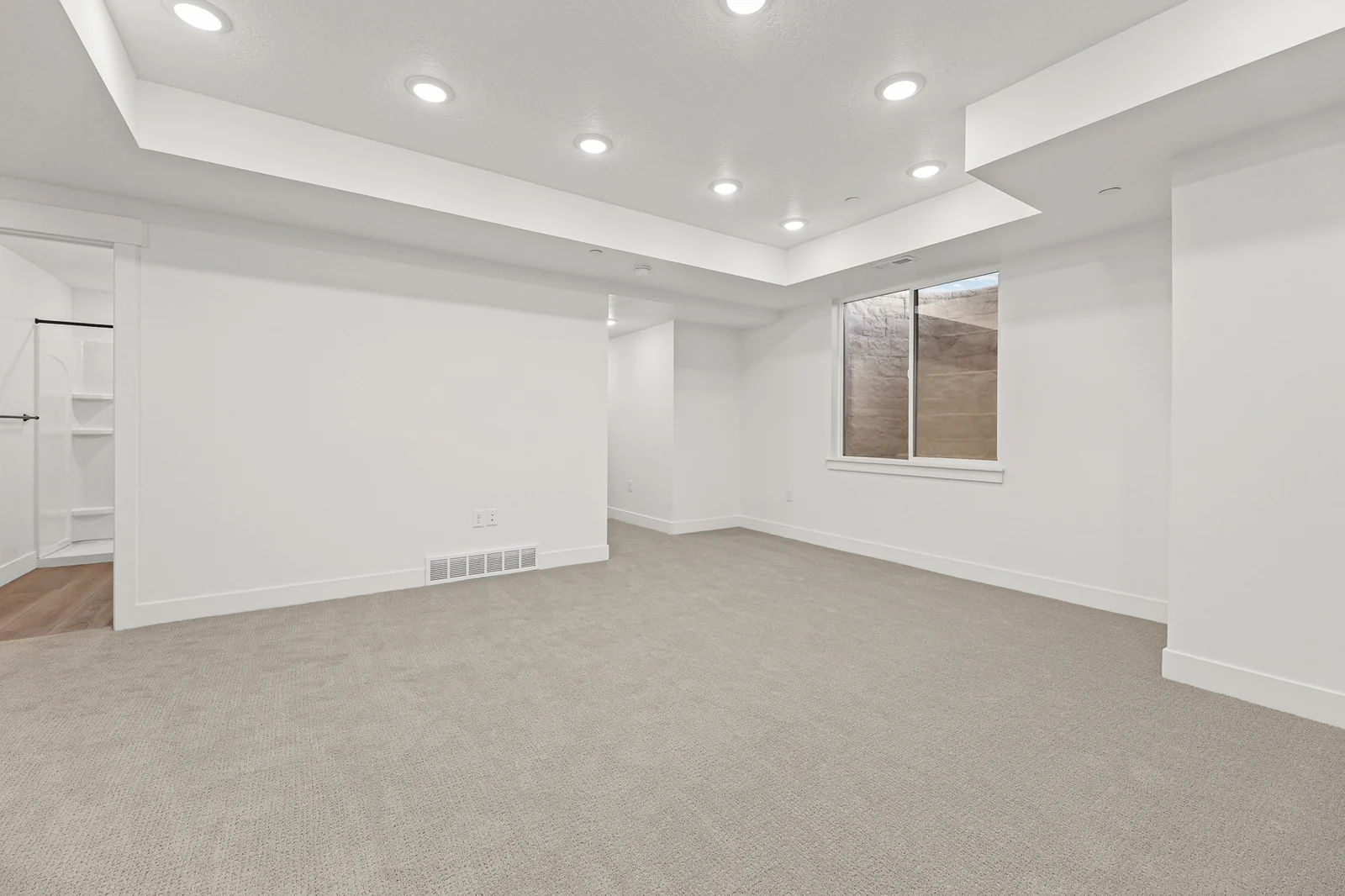 Open carpeted lower-level recreation area with tray ceiling, recessed lights, a window, and white walls in Hawthorne Modern, Ogden.