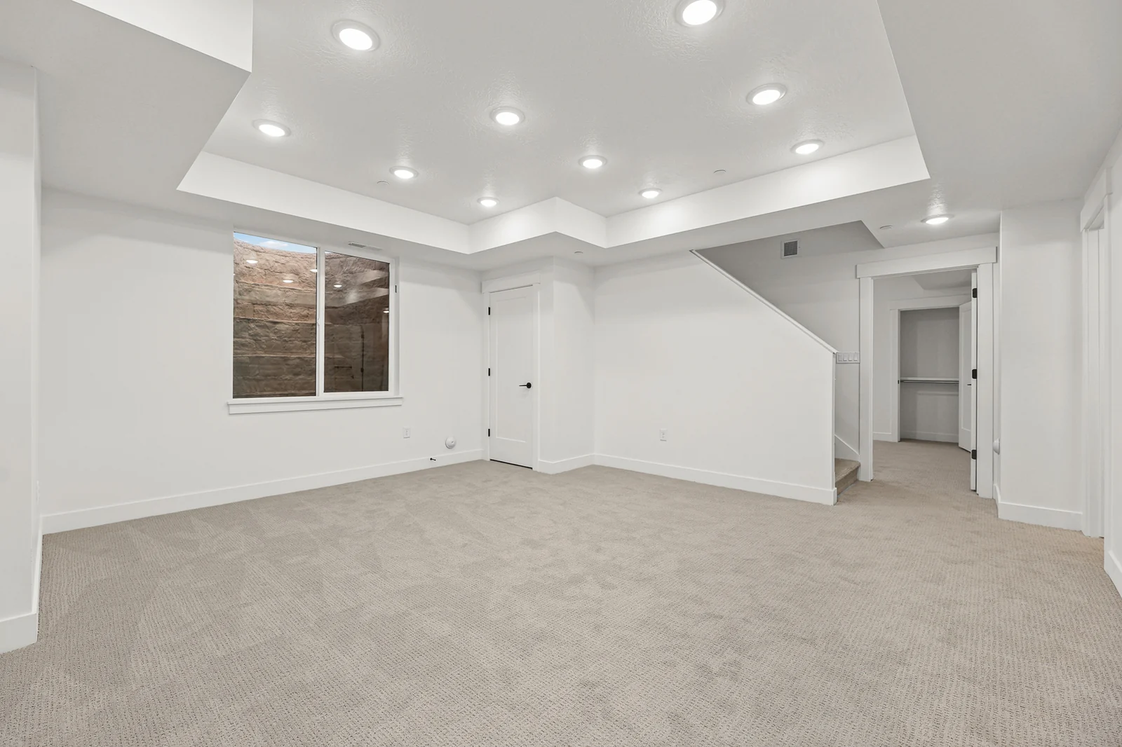 Open lower-level room with carpet, tray ceiling, recessed lights, windows, and a staircase in Hawthorne Modern, Ogden.