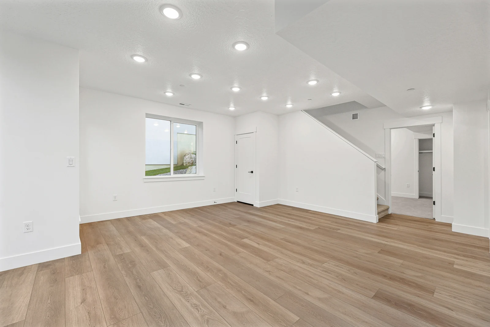 Wide lower-level room shows wood flooring, a window, open areas, a stairway, and white walls in Hawthorne Farmhouse, Ogden.