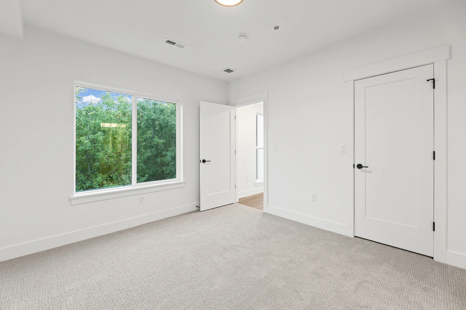 Unfurnished carpeted bedroom includes a wide window, double closet doors, and white trim in Hawthorne Farmhouse, Ogden.