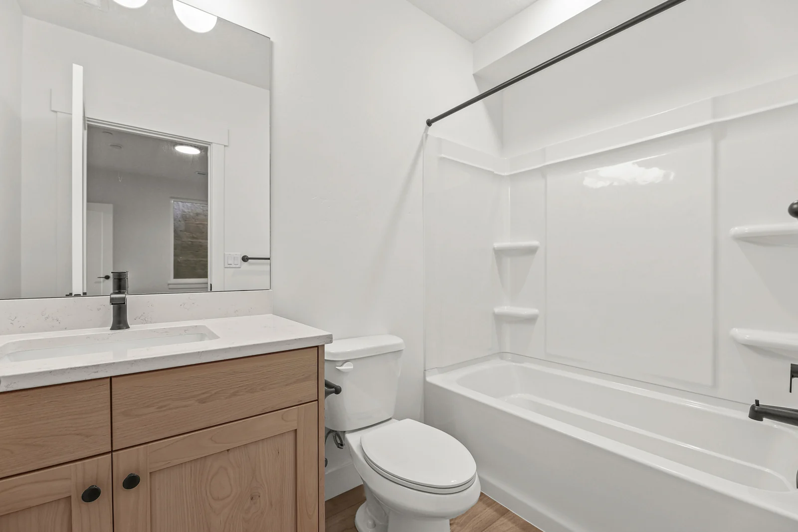 Lower-level bathroom with wood vanity, toilet, white tub-shower surround, black fixtures, and tile floor in Hawthorne Modern, Ogden.