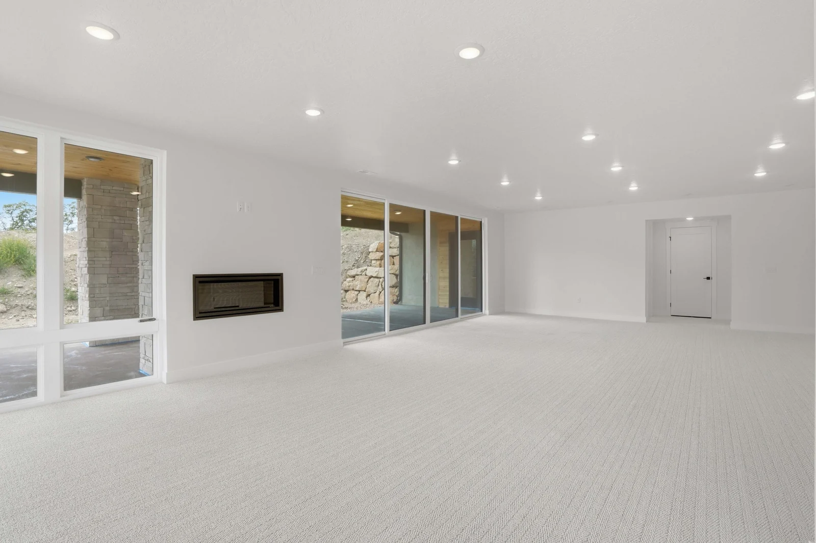 Open lower-level room with pale carpet, recessed lights, a linear fireplace, and wide sliding glass doors in the Denali model, American Fork.