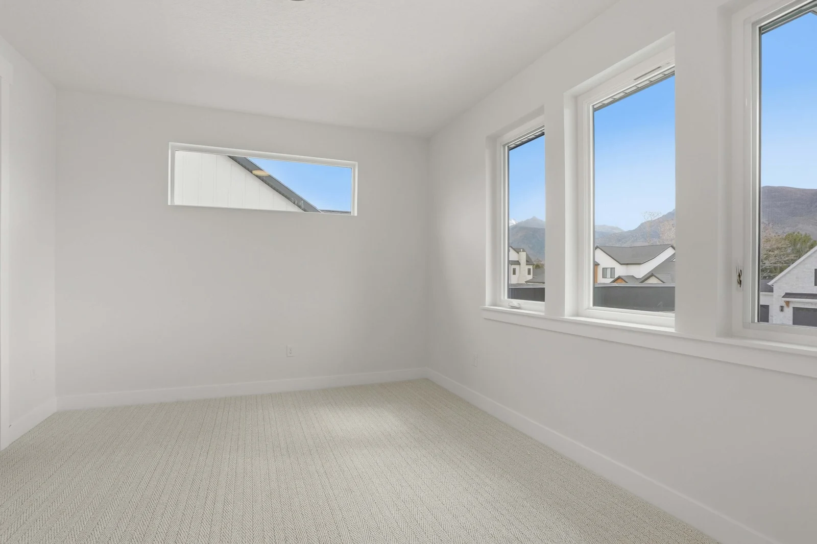 Unfurnished bedroom with pale carpet, a high horizontal window, tall corner windows, and views of nearby rooftops in the Denali model, American Fork.