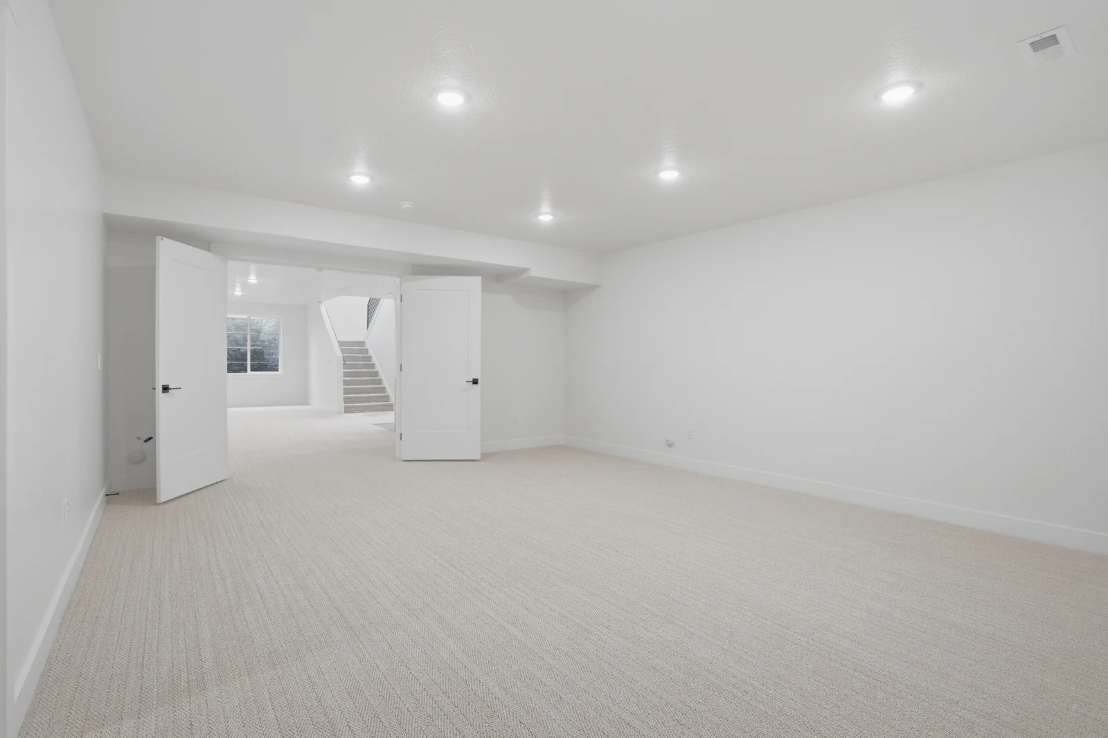 Open lower-level rec room with recessed lights, neutral carpet, double doors, and stairs beyond in Olympic BH08, American Fork.