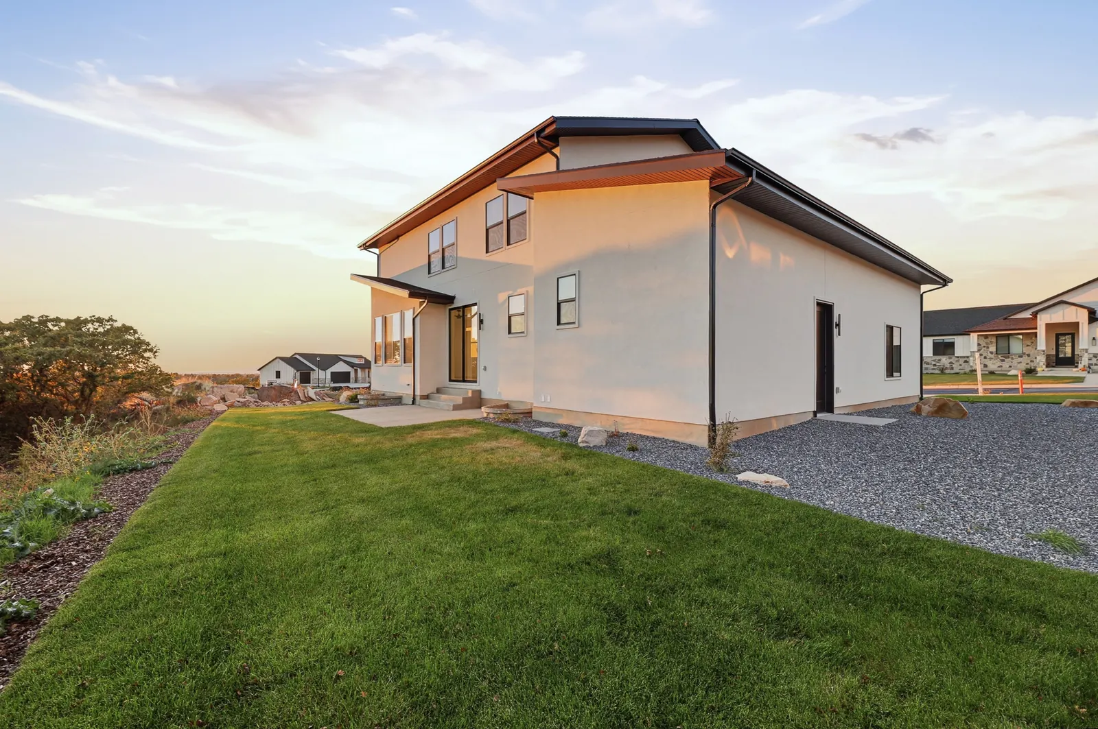 Concrete rear patio beside a lawn, rock border, trees, and a distant sunset view in Hawthorne Modern, Ogden.