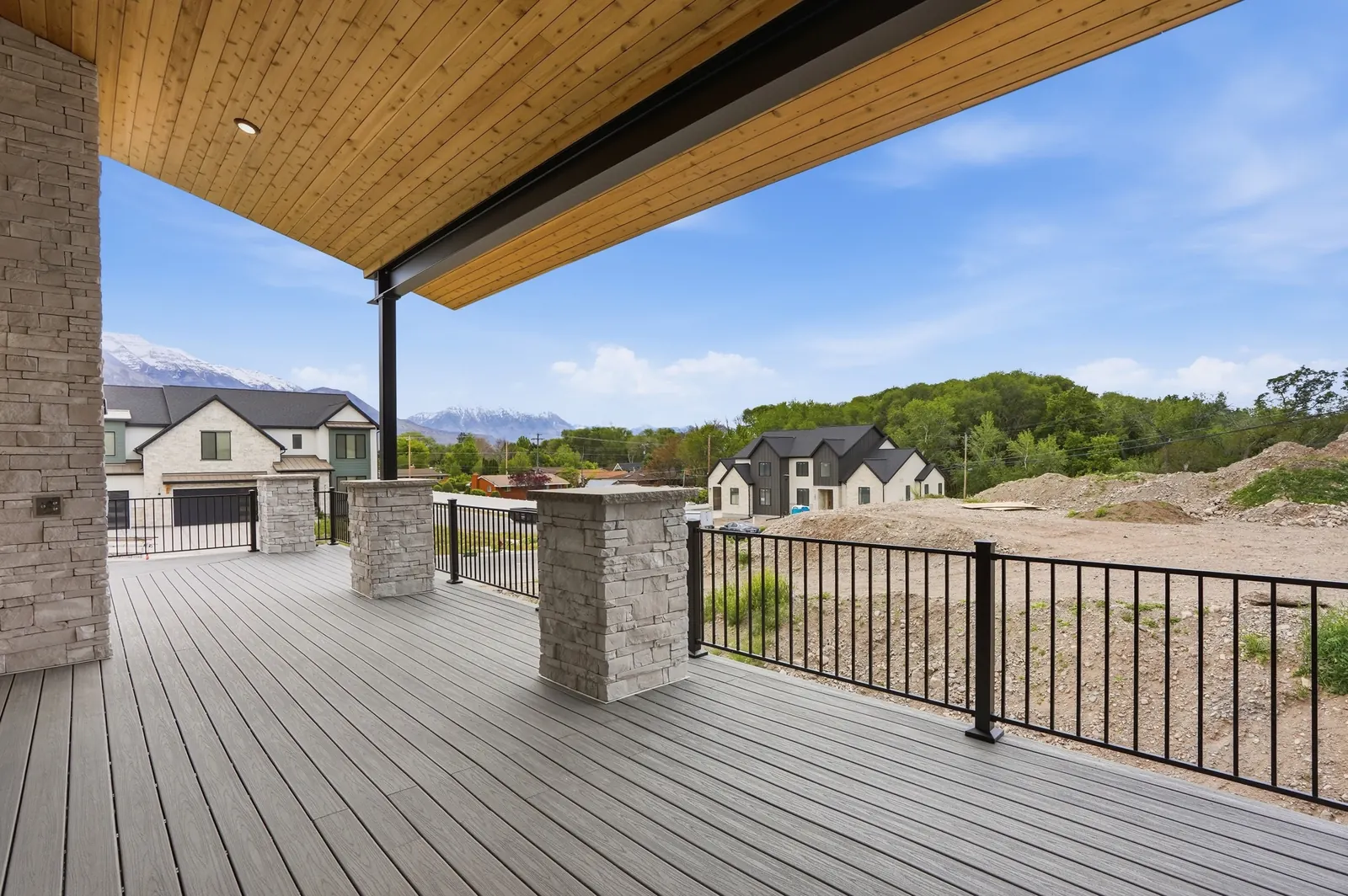 Covered deck with composite flooring, stone columns, black railing, wood ceiling, and views across nearby homes in the Denali model, American Fork.
