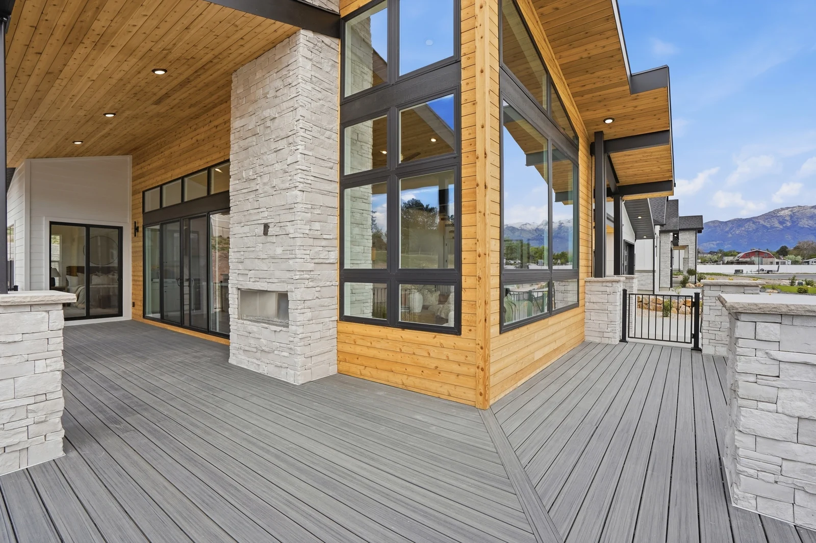 Covered deck beside a stone outdoor fireplace, with wood ceiling, dark trim, glass doors, and black metal railing in the Denali model, American Fork.