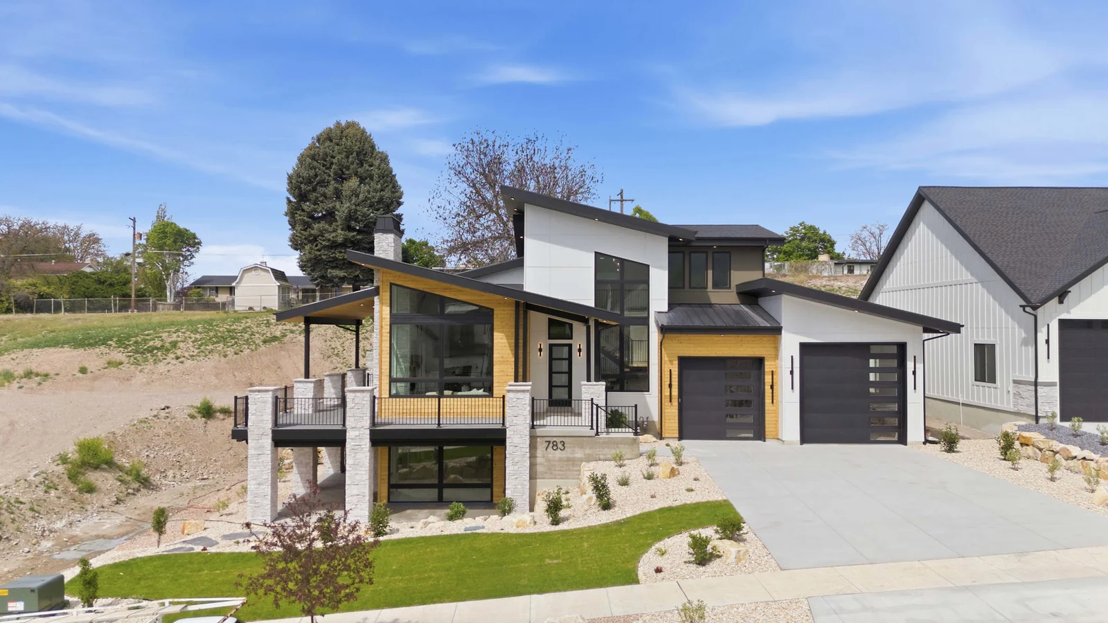 Low aerial view of the modern front exterior, driveway, lawn, garage doors, and neighboring homes in the Denali model, American Fork.