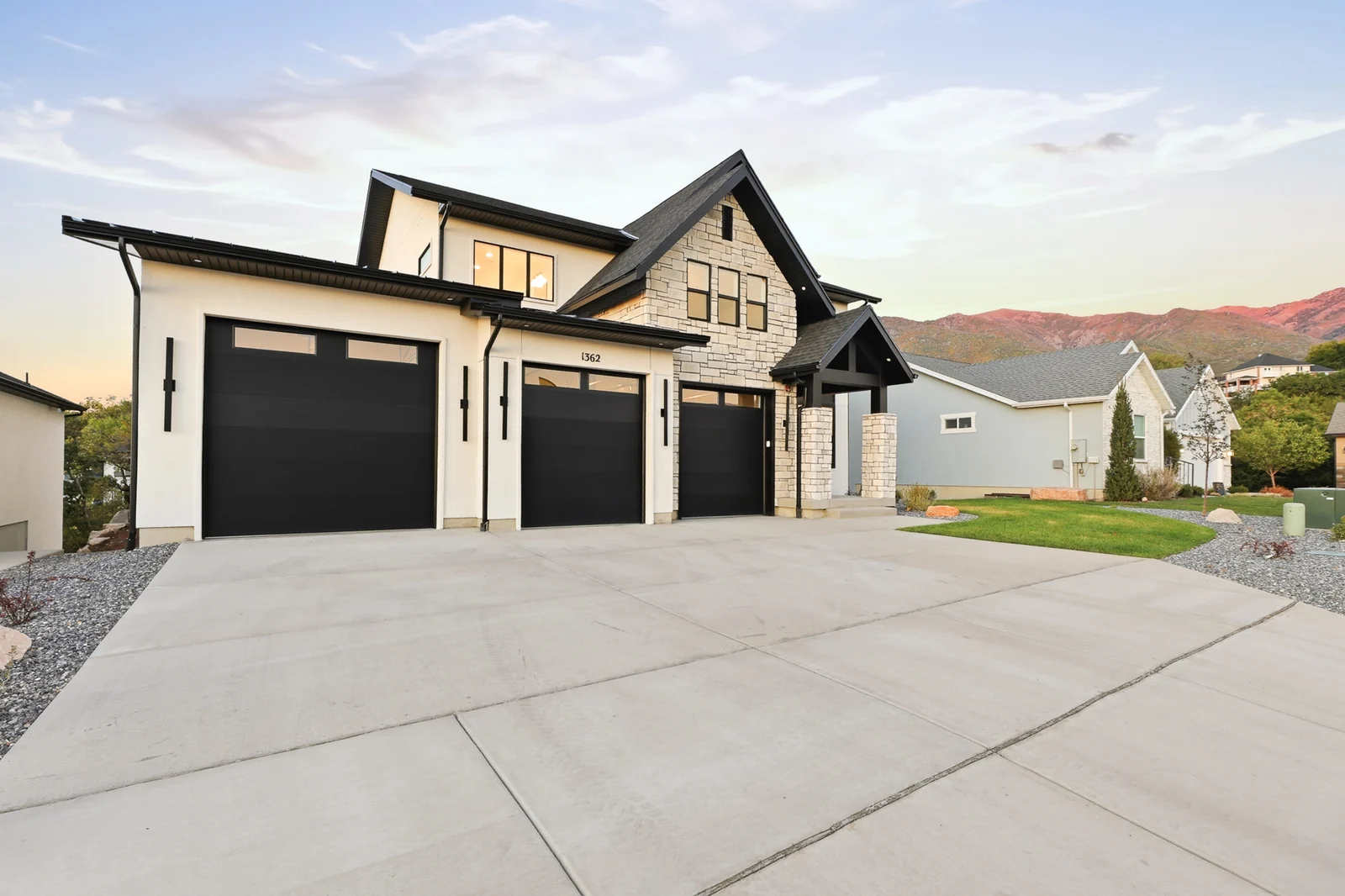 Front exterior shows three dark garage doors, a broad concrete driveway, light facade, and dark gables in Hawthorne Farmhouse, Ogden.