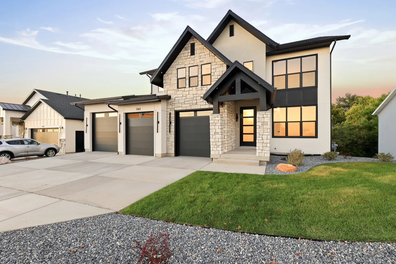 Front exterior view shows dark gables, illuminated windows, garage doors, driveway, lawn, and surrounding homes in Hawthorne Farmhouse, Ogden.