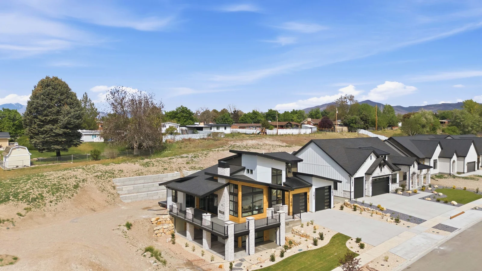 Aerial side view of the modern exterior, covered deck, lawn, neighboring homes, and open ground beside the property in the Denali model, American Fork.