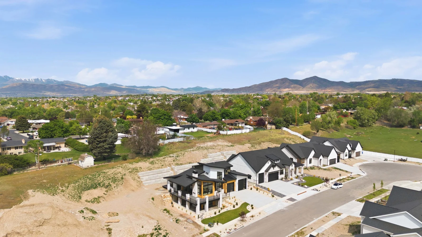 Wide aerial view showing the home, nearby residences, open lots, tree cover, and foothills in the surrounding setting of the Denali model, American Fork.