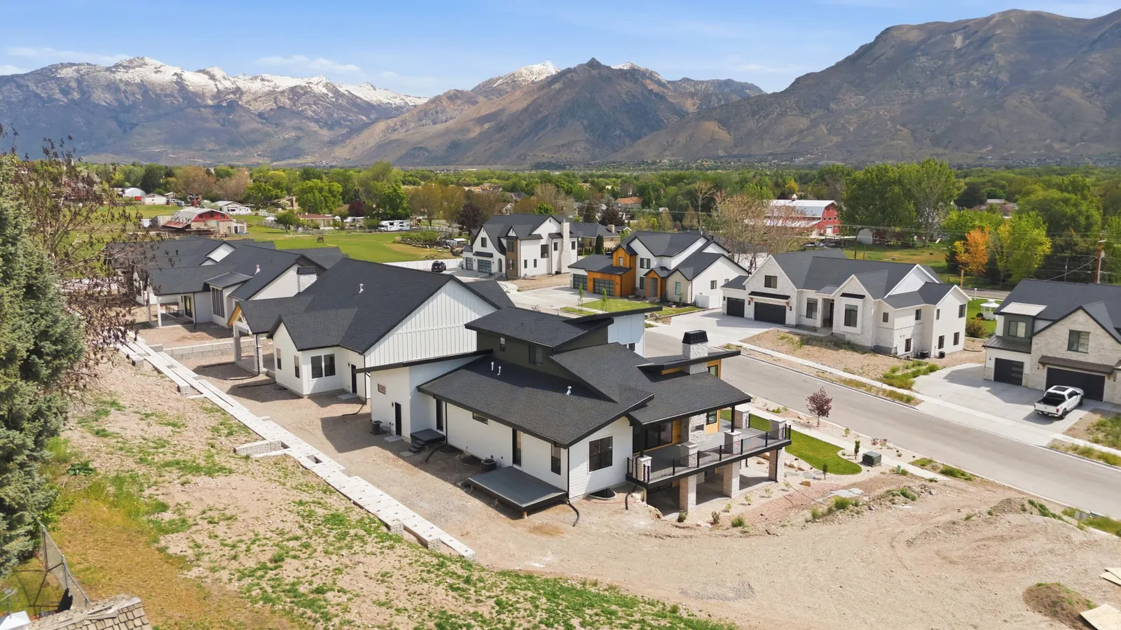 Wide aerial view over the home, neighborhood roofs, fields, tree canopy, and mountain backdrop in the Denali model, American Fork.