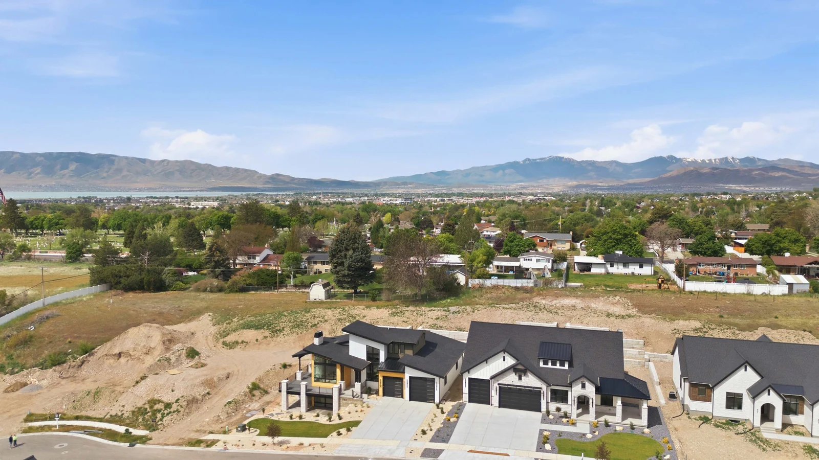 Elevated aerial view across the home, nearby neighborhood, green tree canopy, open fields, and mountain ridgeline in the Denali model, American Fork.