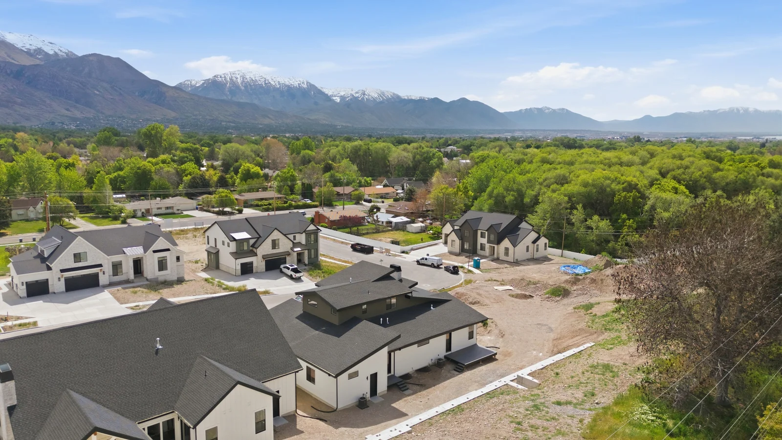 Wide aerial outlook over the home, neighborhood streets, treetops, distant fields, and layered mountains in the Denali model, American Fork.