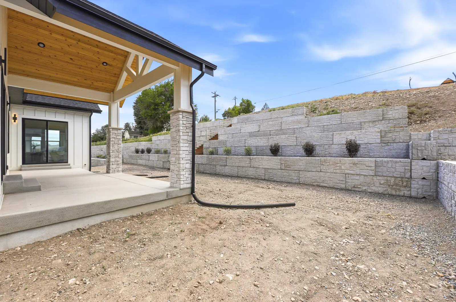 Covered rear patio with timber roof, stone columns, concrete surface, and retaining wall beyond in Olympic BH08, American Fork.