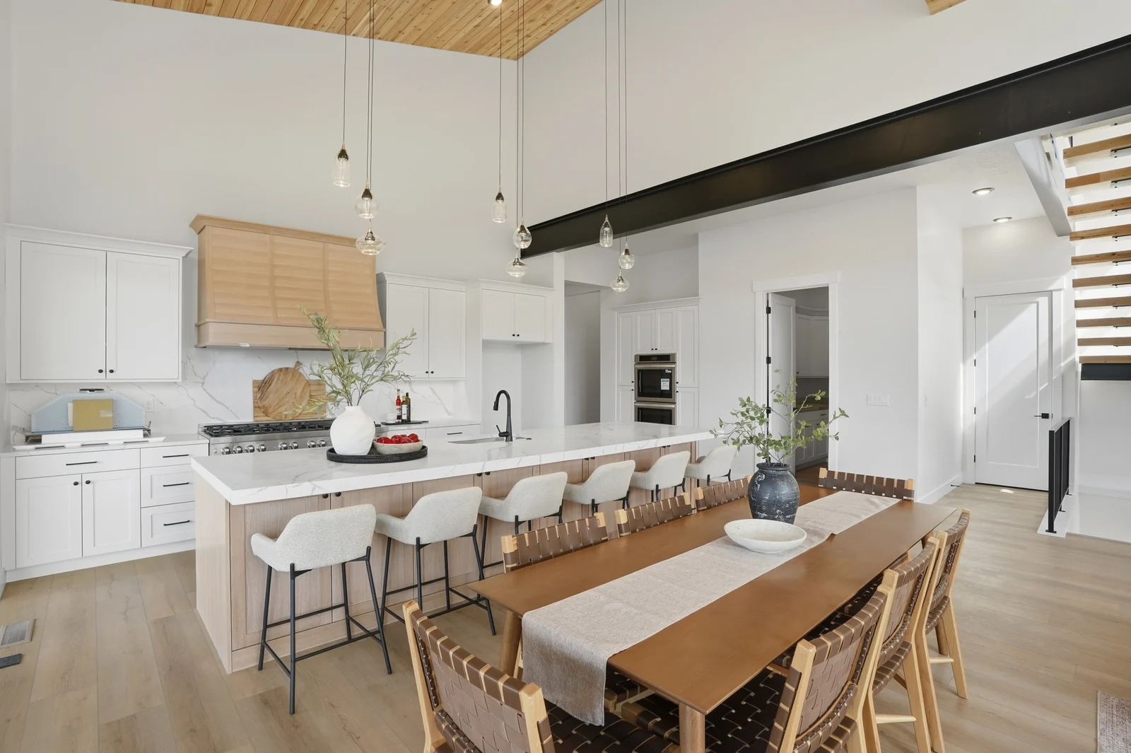 Wide great room and kitchen view with a stone fireplace, leather seating, island stools, wood ceiling, and open stair in the Denali model, American Fork.