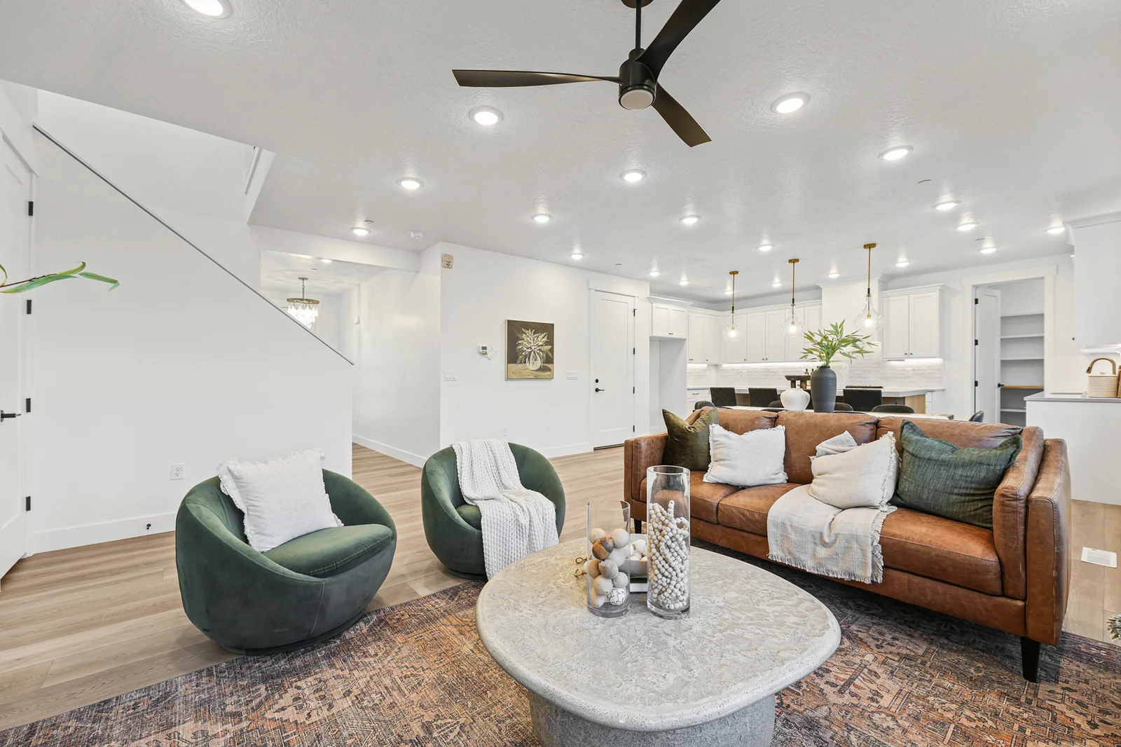 Staged living room features curved chairs, leather sofa, round table, stairway, and recessed lighting in Hawthorne Farmhouse, Ogden.