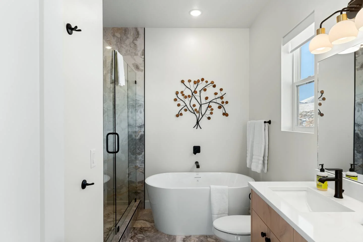 Bathroom with a freestanding soaking tub, glass shower, single vanity, tree wall decor, and white finishes — Denali, Bear Lake.