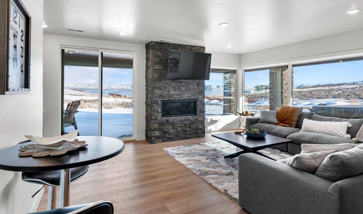 Lower-level living room with sectional seating, stone fireplace, round table, expansive windows, and mountain-facing views — Denali, Bear Lake.