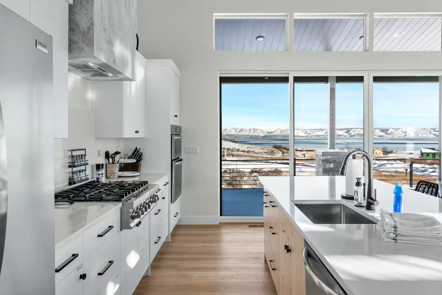 Kitchen with white cabinetry, stainless appliances, island sink, black countertop, wood ceiling detail, and wide windows — Denali, Bear Lake.