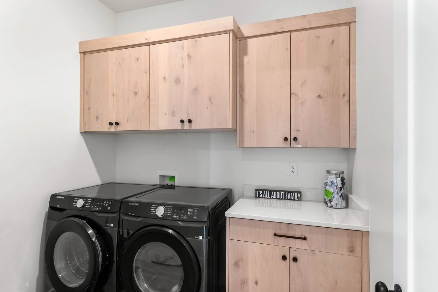 Laundry room with side-by-side machines, pale wood upper cabinets, lower cabinetry, white walls, and black hardware — Denali, Bear Lake.