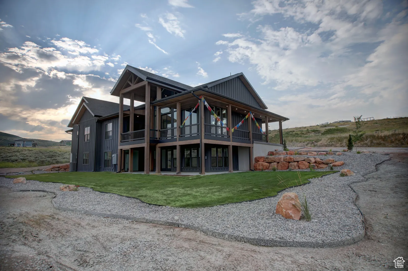 Rear angled exterior view showing the covered deck, dark siding, lawn, gravel drive, and open terrain in Pinnacle, Bear Lake.